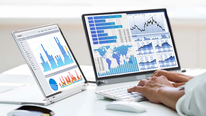 Hands typing on computer keyboard with two screens showing charts and graphs